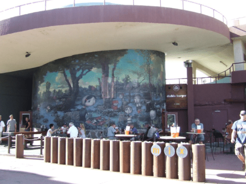 A colorful mural on a round building, with outdoor seating and people enjoying food at a casual eatery.