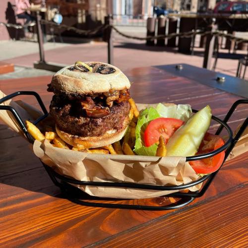 A delicious burger with bacon, lettuce, tomato, and pickles, served with fries in a black basket on a wooden table.
