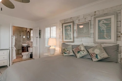 Cozy bedroom with a large bed, decorative pillows, and a bathroom visible in the background. Nautical-themed decor.