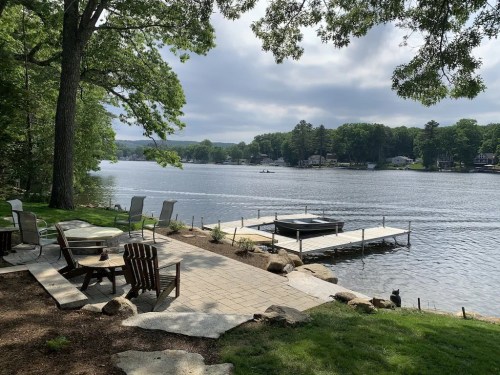 A serene lakeside view with a dock, chairs, and lush greenery under a cloudy sky.