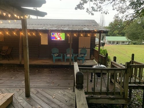 A rustic cabin with string lights, a TV on the wall, and blue chairs on a wooden deck overlooking a grassy area.