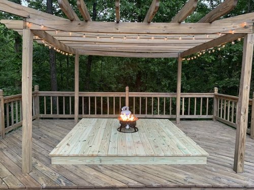 A wooden deck with a fire pit in the center, surrounded by trees and string lights overhead.