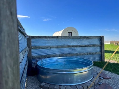 A metal water trough sits on gravel, with a covered wagon in the background under a clear blue sky.