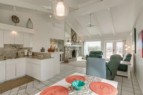 Bright, modern living space with a kitchen, dining area, and cozy seating by a stone fireplace and large windows.