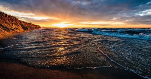 Sunset over a beach with waves gently lapping at the shore, illuminated by warm golden light and dramatic clouds.