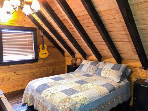 Cozy attic bedroom with a quilted bed, wooden walls, and a guitar hanging on the wall. Warm lighting from a chandelier.