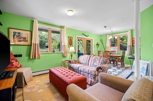 Cozy living room with green walls, floral couch, red ottoman, and dining area with wooden table and chairs.