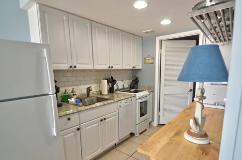 A small kitchen with white cabinets, a sink, stove, and a wooden table with a blue lamp.