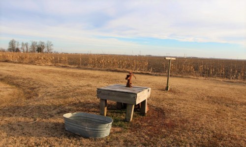 A rustic water pump on a wooden platform, surrounded by a barren field and a metal tub under a clear sky.