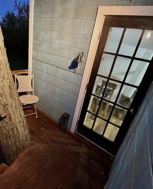A cozy porch area with a chair, lantern, and a door leading into a warmly lit interior.
