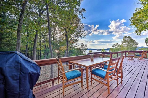 A wooden deck with a table and chairs, surrounded by trees and a view of the water under a blue sky with clouds.