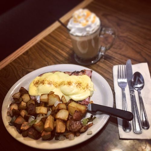 A plate of eggs Benedict with potatoes, alongside a coffee topped with whipped cream, on a wooden table.