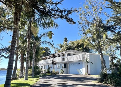 A modern two-story house surrounded by palm trees, located by a serene body of water under a clear blue sky.