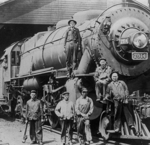 Black and white photo of six men posing with a steam locomotive, one standing on the front, in a train yard.