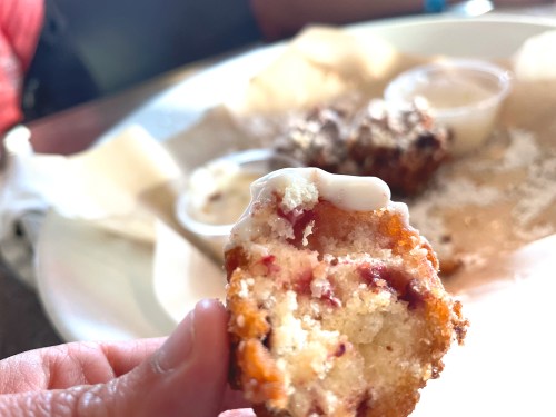 A close-up of a hand holding a bite of a muffin with a creamy topping, with more muffins and dipping sauce in the background.