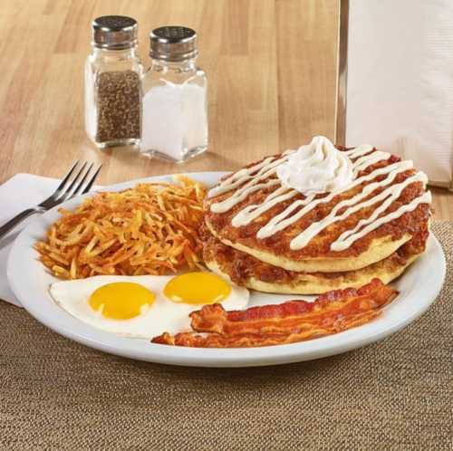 A plate of breakfast featuring pancakes with whipped cream, eggs, bacon, and hash browns on a wooden table.