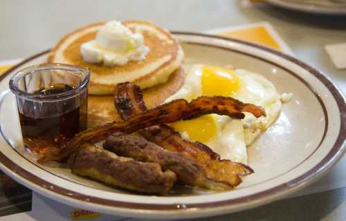 A plate of breakfast featuring pancakes with butter, two eggs, bacon, sausage, and a small cup of syrup.