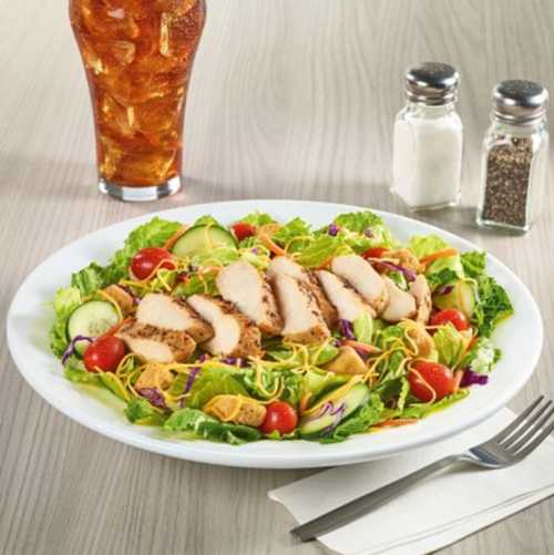 A colorful salad with sliced chicken, cherry tomatoes, cucumbers, and mixed greens, served with a drink and condiments.