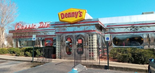 A shiny, retro-style Denny's diner with a bright yellow sign, surrounded by greenery and a clear blue sky.