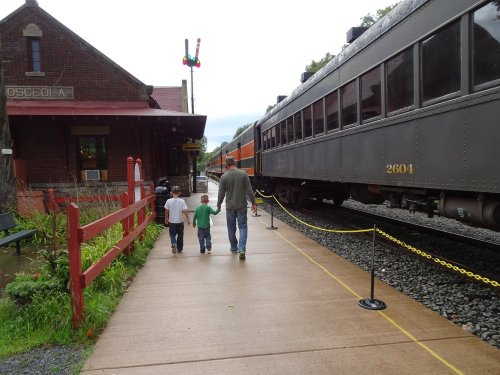 Two children and an adult walk along a sidewalk beside a train station, with vintage trains parked nearby.