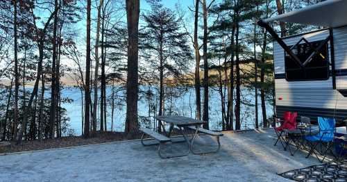 A serene lakeside campsite with a picnic table, chairs, and trees surrounding a tranquil water view.