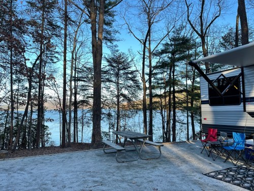 A serene campsite by a lake, featuring a picnic table, chairs, and trees in the background under a clear blue sky.
