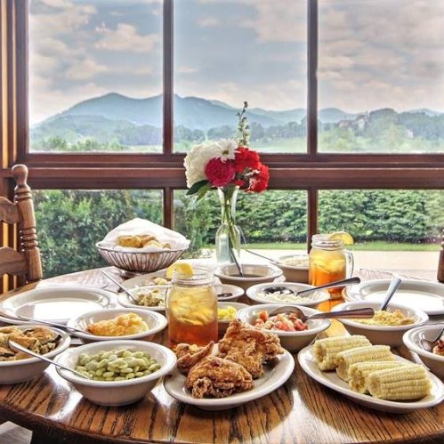 A table filled with various dishes, including fried chicken, corn, and sides, with a scenic mountain view outside.