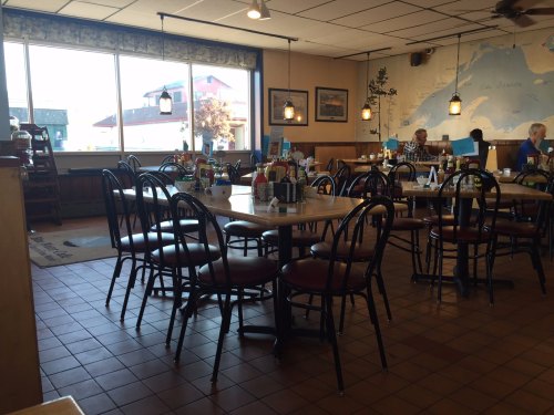 A cozy restaurant interior with empty tables, chairs, and large windows showing a view outside.