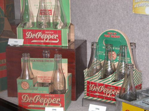 Vintage Dr. Pepper bottles and packaging displayed in a museum setting, showcasing designs from 1939 and 1946.