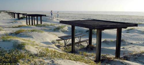 Two people walk along a sandy beach with wooden structures and picnic tables in the foreground.