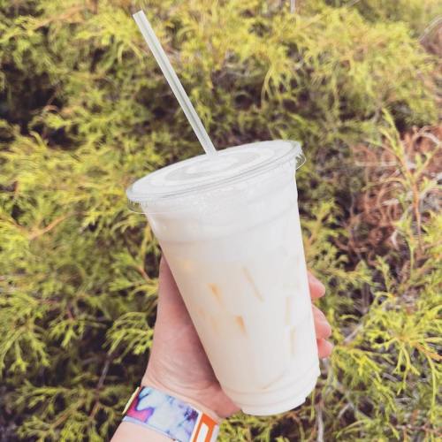A hand holding a clear cup of iced drink with a straw, set against a backdrop of green foliage.