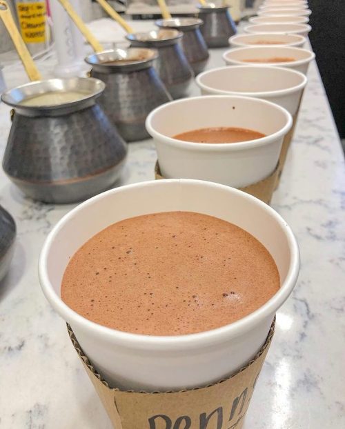 A row of white cups filled with frothy chocolate drink on a marble countertop, with metal pots in the background.