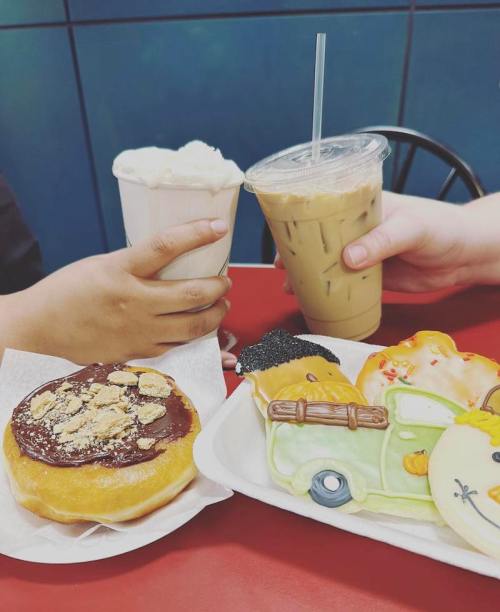 Two hands holding drinks, with a donut and decorated cookies on a table.