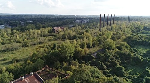 Aerial view of a lush, overgrown landscape with remnants of industrial structures and smokestacks in the distance.
