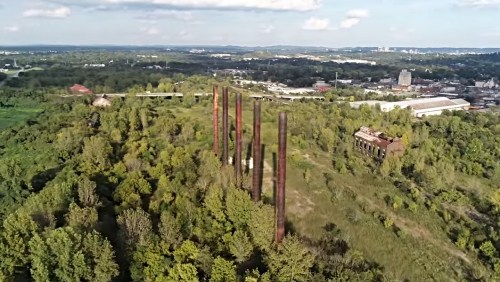 Aerial view of overgrown land with tall, rusty smokestacks and remnants of industrial buildings in the background.