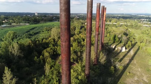 Aerial view of tall, rusty smokestacks surrounded by lush greenery and trees under a clear blue sky.