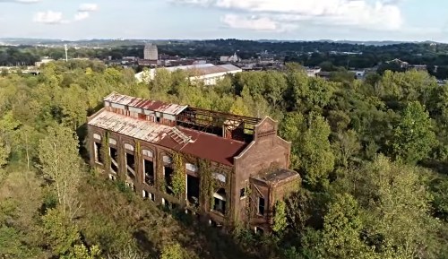 An abandoned, overgrown brick building surrounded by trees, with a city skyline visible in the background.