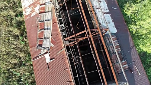 Aerial view of a dilapidated building with a rusted roof and exposed structure, surrounded by overgrown vegetation.
