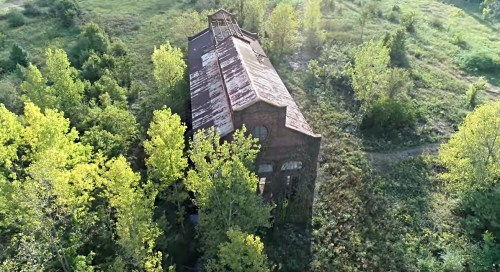 Aerial view of an abandoned brick building surrounded by lush green trees and overgrown vegetation.