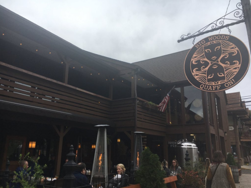 A rustic restaurant exterior with a sign reading "Big Woods Quaff On," featuring outdoor seating and cloudy skies.