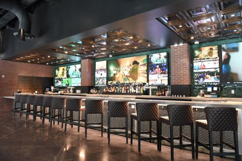 A modern bar with a sleek design, featuring a long counter, high stools, and multiple screens displaying various programs.