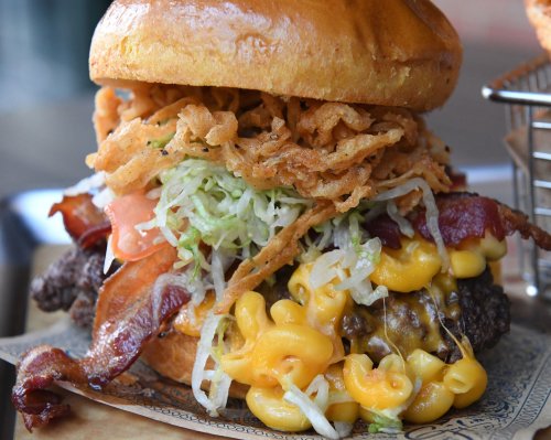A towering burger with bacon, crispy onions, lettuce, tomato, and mac and cheese on a toasted bun.
