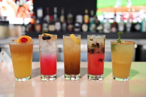 Five colorful cocktails lined up on a bar, featuring garnishes like fruit and herbs, with a blurred background of bottles.