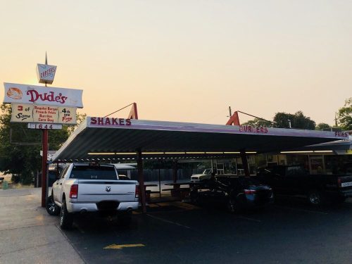 A retro diner named "Dude's" with a sign for shakes and burgers, featuring parked cars under a covered area.