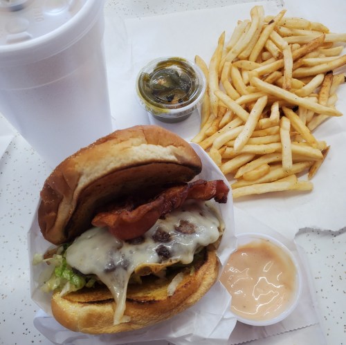 A burger with bacon and cheese, fries, pickles, and two dipping sauces, alongside a large drink.