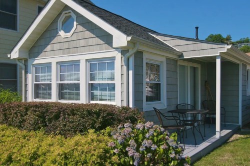 A cozy gray house with a porch, featuring large windows and a small garden with blooming flowers.