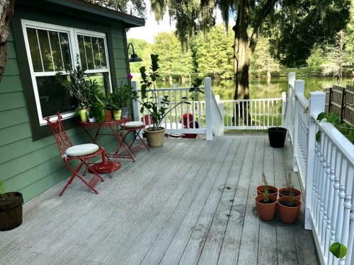 A serene deck with red chairs, potted plants, and a view of a calm lake surrounded by trees.