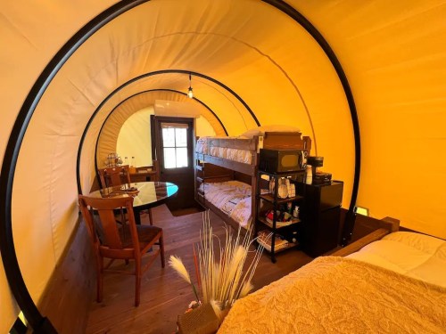 Cozy interior of a glamping tent featuring bunk beds, a dining table, and rustic decor. Warm yellow tones throughout.