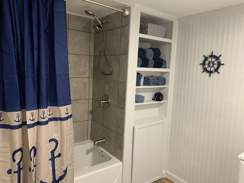 A bathroom featuring a shower with a nautical-themed curtain, shelves of towels, and a ship's wheel decoration.