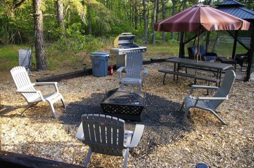 A cozy outdoor space with a fire pit, chairs, a grill, and a gazebo surrounded by trees and wood chips.